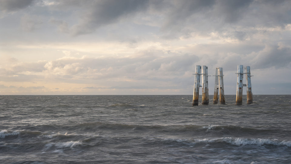 The same ocean after a storm: lingering swells, broken clouds, and the distant structures, now weathered, catching an early sunrise.