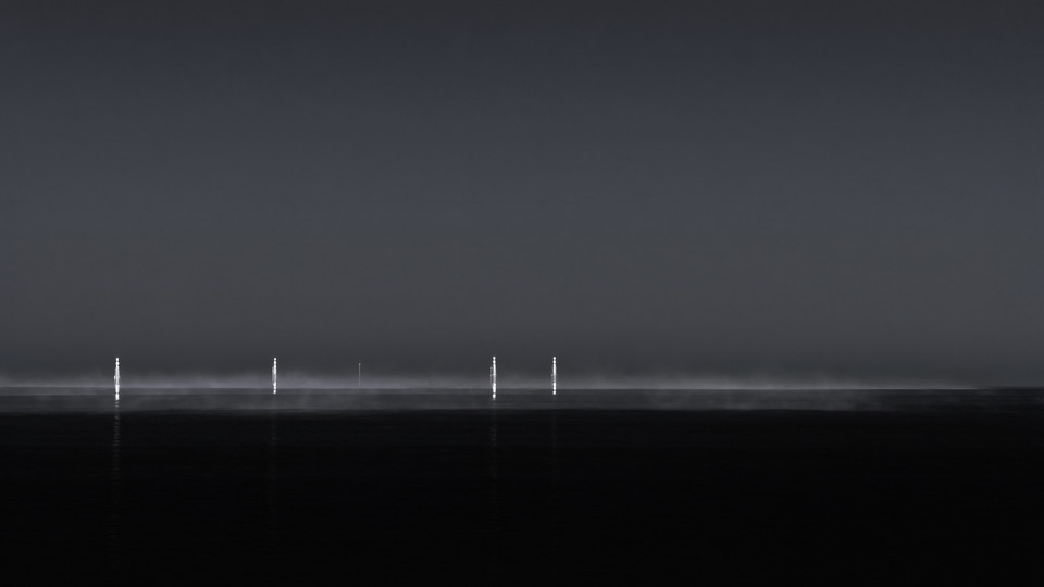 A quiet black ocean at night, with distant silver structures standing far out at sea in thin fog.